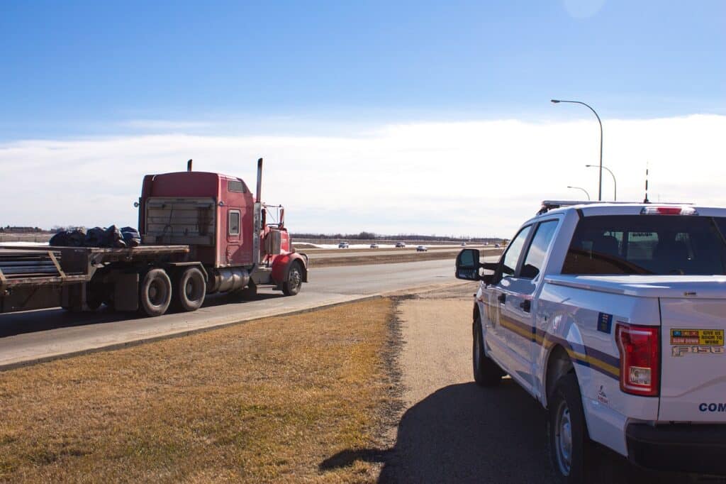A police car sits in an off ramp while a semi truck-trailer drives on main highway