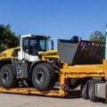 A white and yellow loader on a low loader trailer attached to a yellow and blue semi truck