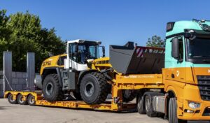A white and yellow loader on a low loader trailer attached to a yellow and blue semi truck