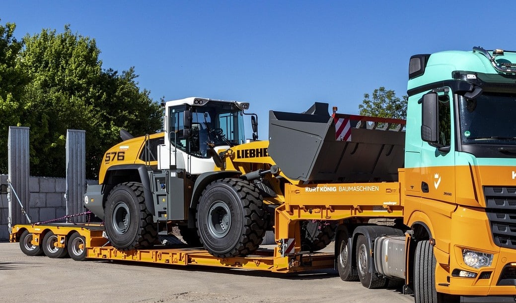 A white and yellow loader on a low loader trailer attached to a yellow and blue semi truck